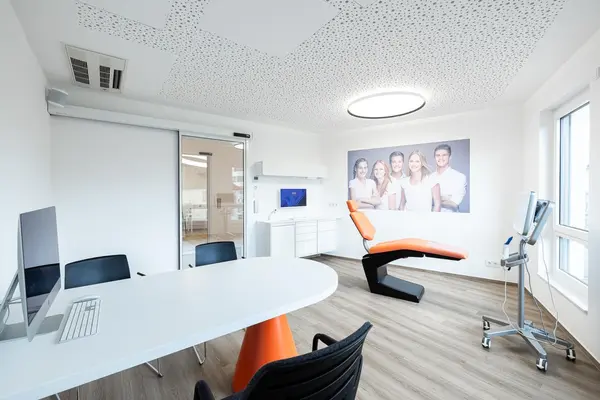 Modernes Behandlungszimmer mit orangefarbener Liege, Computer und heller Praxiseinrichtung.