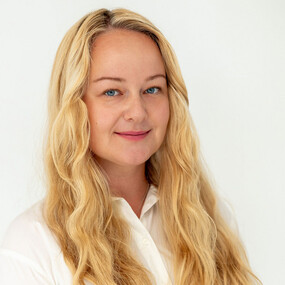 Porträt einer lächelnden Frau mit langen blonden Haaren vor neutralem Hintergrund.