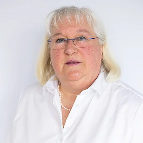 Porträt einer älteren Frau mit Brille und weißer Bluse vor neutralem Hintergrund.
