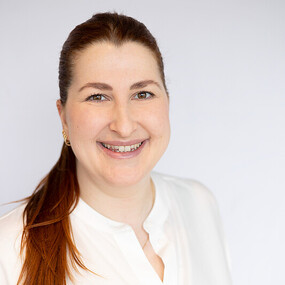Porträt einer lächelnden Frau mit braunen Haaren im weißen Hemd vor hellem Hintergrund.