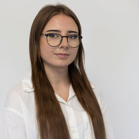 Junge Frau mit langen braunen Haaren und Brille im weißen Hemd vor hellem Hintergrund.