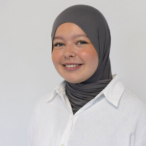 Junge Frau mit grauem Hijab und weißer Bluse lächelt freundlich vor hellem Hintergrund.