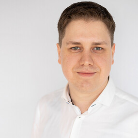 Porträt eines jungen Mannes mit kurzen braunen Haaren und weißem Hemd vor hellem Hintergrund.