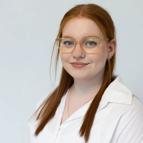 Junge Frau mit Brille und roten Haaren lächelt im weißen Hemd vor hellem Hintergrund.