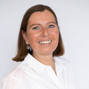 Porträt einer lächelnden Frau mit braunen Haaren und weißer Bluse vor weißem Hintergrund.