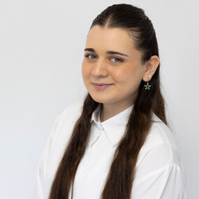 Junge Frau mit langen braunen Haaren und weißer Bluse vor neutralem Hintergrund.